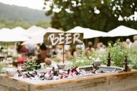 a rustic wedding bar of reclaimed wood, with greenery around, with ice and lots of bottles inside is a very cool and creative idea