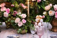 a sophisticated garden wedding table setting with greenery and pink rose centerpieces, printed plates, blush glasses and a tablecloth