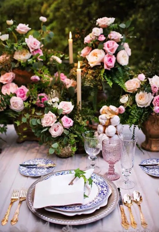 a sophisticated garden wedding table setting with greenery and pink rose centerpieces, printed plates, blush glasses and a tablecloth