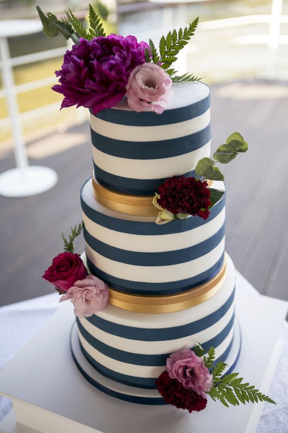 a striped navy and white wedding cake with gold stripes, pink, burgundy and fuchsia blooms and greenery for a summer wedding
