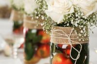 a wedding centerpiece of a jar filled with apples and greenery, with white roses and baby’s breath, a burlap wrap for the jar