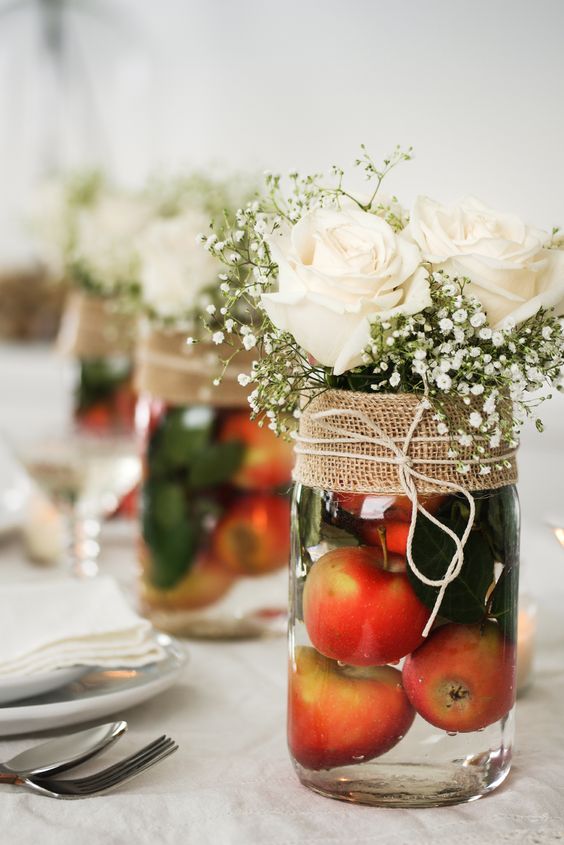 a wedding centerpiece of a jar filled with apples and greenery, with white roses and baby's breath, a burlap wrap for the jar