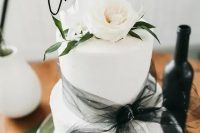 a white buttercream wedding cake with a black tulle bow, with white roses and greenery on top and a black calligraphy topper for a black and white wedding