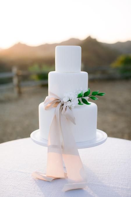 a white buttercream wedding cake with white blooms, greenery and a greige ribbon bow is a lovely and chic idea for a modern wedding