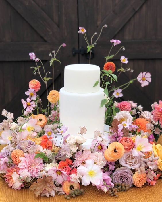 a white wedding cake surrounded with lush orange, yellow and lilac blooms and greenery all over are great for a garden wedding