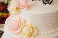 a white wedding cake with neutral and pink sugar roses and a bike cake topper is a stylish idea for a spring or summer wedding