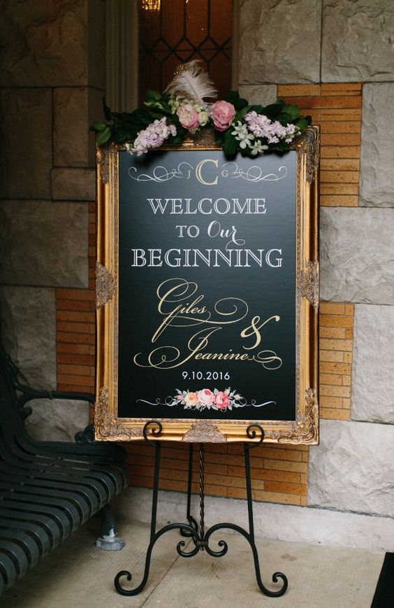 an elegant chalkboard sign in a very refined and beautiful gilded frame with floral arrangements, greenery and feathers