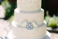 an elegant white wedding cake with lace and plain tiers, with a sugar ribbon bow and an embellished brooch is amazing for a vintage wedding