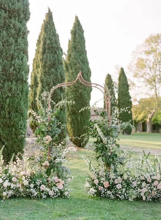 an enchanting garden wedding arch of a whimsy shape, with greenery and lots of blush blooms climbing up the arch