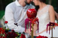 exquisite jewel-toned fall wedding decor with red blooms, berries, greenery and apples, a tall gold stand with berries and an apple with callgiraphy