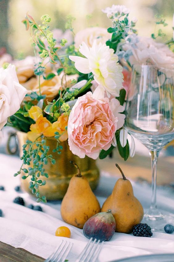 a beautiful and delicate wedding centerpiece of a gold vase with pastel and neutral blooms and greenery, pears, figs and blackberries on the table