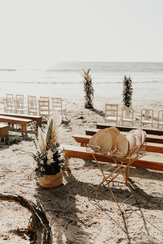 a beautiful beach wedding ceremony space with white chairs and stained benches, white blooms, greenery and pampas grass
