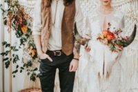 a boho groom’s look with a white shirt, a bolo tie, a brown leather vest, black jeans, brown shoes and a camel hat