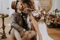 a boho groom’s outfit in earthy tones with tan pants, brown shoes, a black shirt and a brown leather jacket plus a brown hat