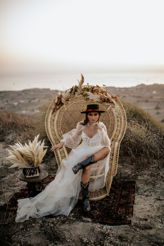 a bold boho bridal look with a semi sheer tulle wedding dress with a deep neckline, black embroidered cowgirl boots, a black flower embellished hat