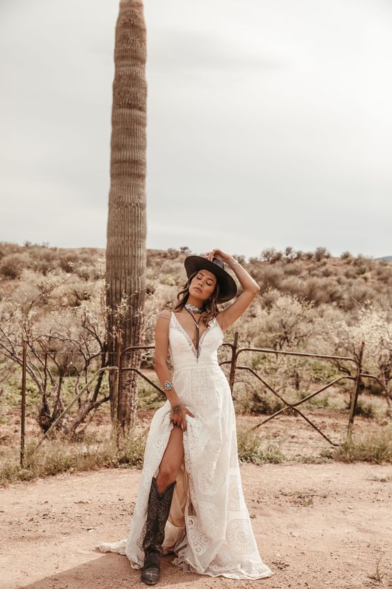 a bold boho wedding look with a lace A-line wedding dress with a plunging neckline, a brown hat, brown cowgirl boots with patterns and statement accessories