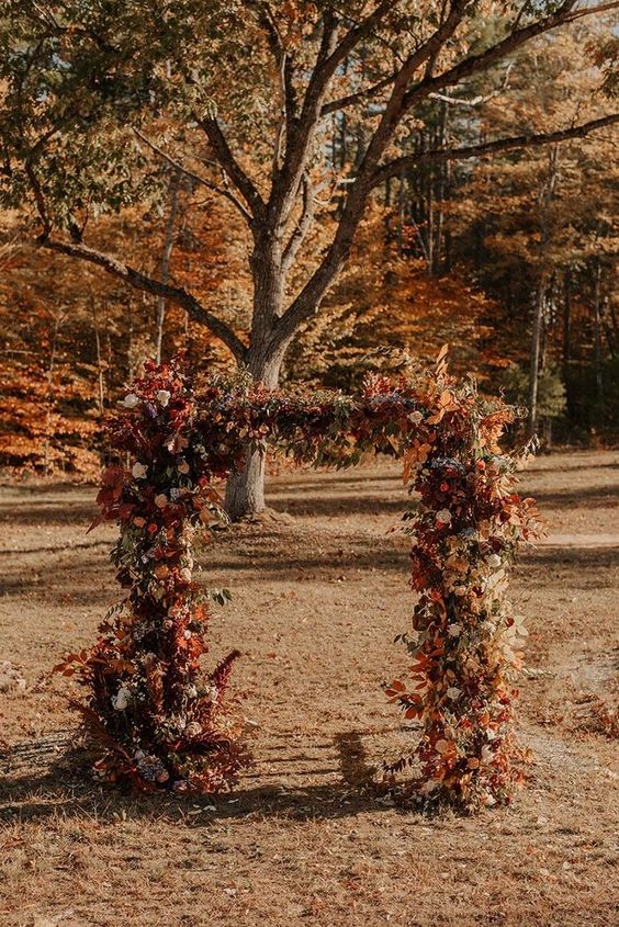 a bold fall wedding arch decorated with neutral and bright foliage, bold blooms and thistles is a cool idea for a fall wedding