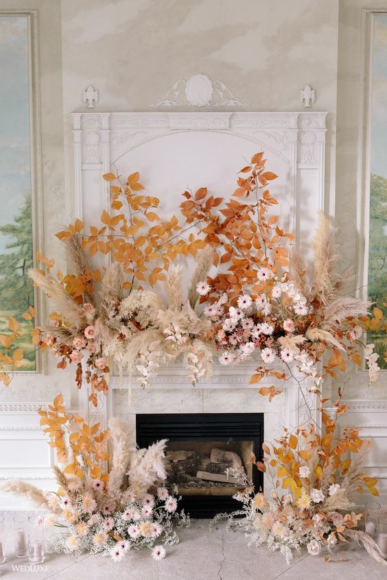 a bright fall wedding backdrop of a fireplace, bold fall leaves, blush and pink blooms and dried flowers plus pampas grass