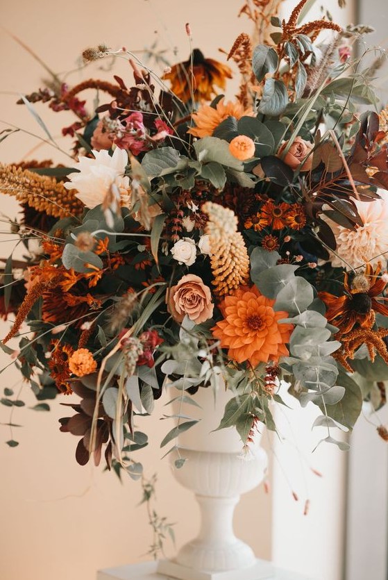 a bright fall wedding centerpiece with a white vintage urn, greenery, lots of blooms in orange, blush, rust and coffee shades