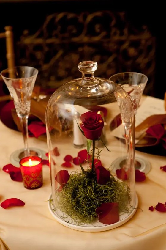 a cloche with a red roses and petals and some grass inside plus candles in red and some petals on the table for a Valentine centerpiece