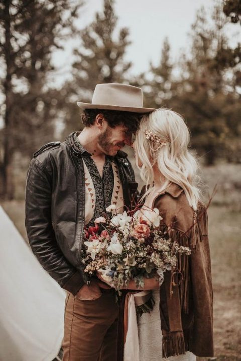 a cool boho groom's outfit with brown pants, a printed shirt, a neutral waistcoat, a black leather jacket and a white hat