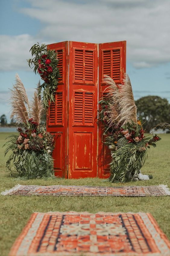 a creative boho wedding backdrop of shabby chic red doors with shutters, greenery, burgundy blooms and pampas grass
