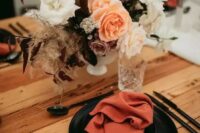 a dramatic fall wedding tablescape with rust napkins, black plates, a pretty bold floral centerpiece, black cutlery and neutral candles