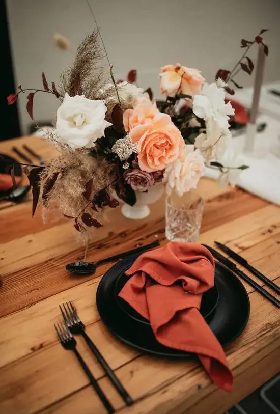 a dramatic fall wedding tablescape with rust napkins, black plates, a pretty bold floral centerpiece, black cutlery and neutral candles