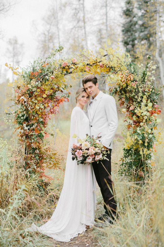 a fall wedding arch covered with greenery and bold fall foliage is a stunning idea for a fall wedding