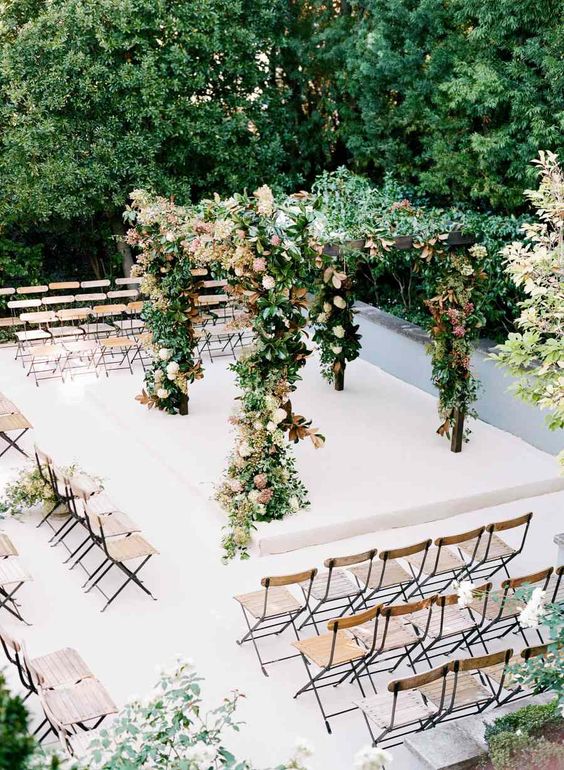 a fall wedding arch covered with greenery, fall blooms and leaves, simple wooden folding chairs all around the space