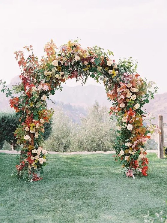 a fall wedding arch decorated with greenery, blush blooms, bold fall leaves and mustard flowers is a cool idea for the fall