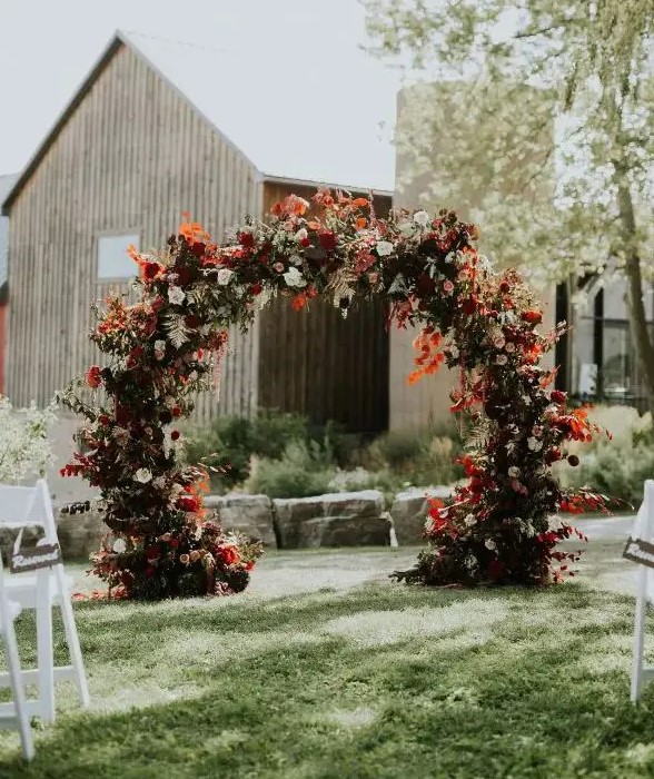 a fantastic round fall wedding arch with blush, burgundy and red blooms, greenery and colorful fall leaves and foliage is amazing