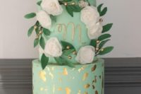 a glam mint and gold flake wedding cake with a monogram, fake blooms and foliage on a chic stand
