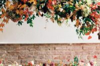 a gorgeous overhead fall wedding installation with greenery and bold fall leaves and a matching arrangement with blooms on the table