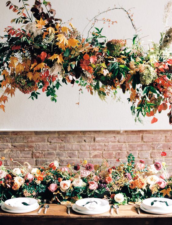 a gorgeous overhead fall wedding installation with greenery and bold fall leaves and a matching arrangement with blooms on the table