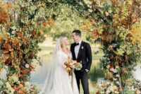 a jaw-dropping fall wedding arch coverd with greenery, white and orange blooms and bold fall leaves is an amazing idea