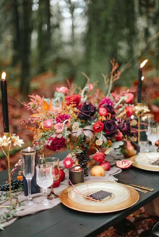a jewel tone wedding centerpiece with lots of greenery, red, pink and purple blooms and grapes, candles and apples on the table