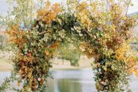 a lush fall wedding arch covered with leaves, green hydrangeas, orange roses and bold fall leaves on top the arch