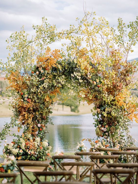 a lush fall wedding arch covered with leaves, green hydrangeas, orange roses and bold fall leaves on top the arch