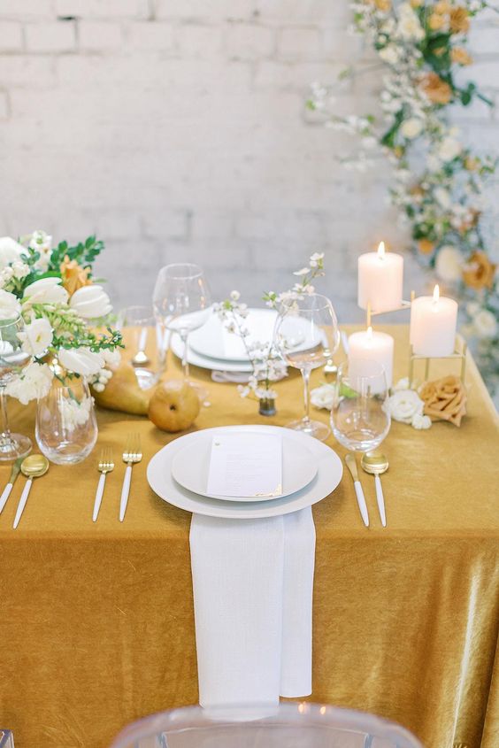 a marigold wedding tablescape with a tablecloth and matching pears, white blooms with greenery, pillar candles and gold and white cutlery