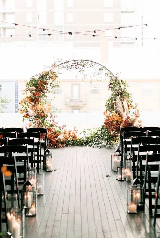 a modern fall wedding ceremony space with a round wedding arch decorated with greenery and bright fall leaves, with candle lanterns lining up the aisle