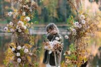 a moody fall wedding arch decorated with blush roses and dahlias, greenery and fall leaves plus a lake view