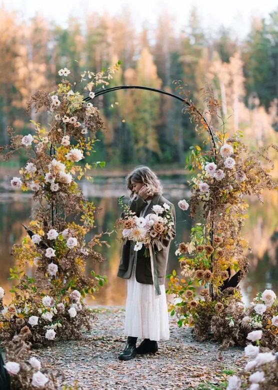 a moody fall wedding arch decorated with blush roses and dahlias, greenery and fall leaves plus a lake view
