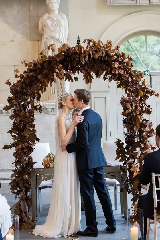 a moody fall wedding arch of dark dried leaves looks refined and rather unusual, it's a creative way to stand out