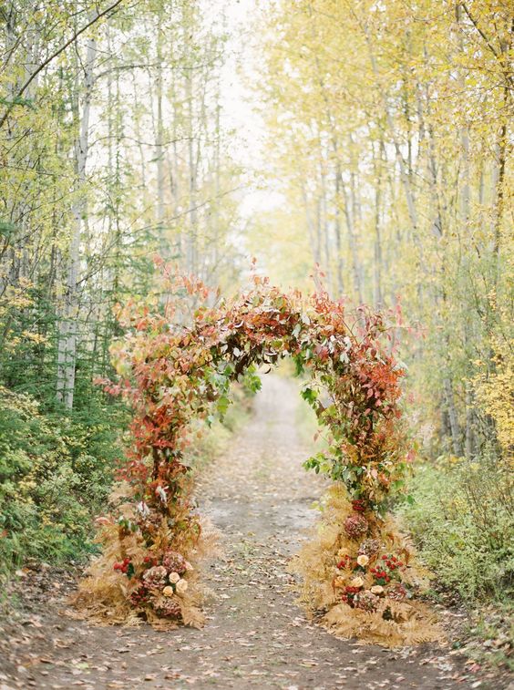 a pretty fall wedding arch covered with greenery, bold leaves and branches, neutral and bold blooms is amazing