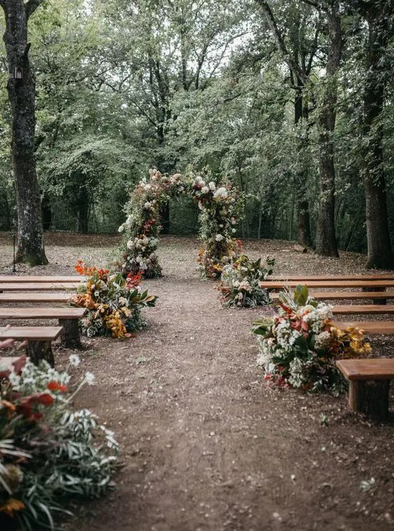 a pretty fall woodland wedding aisle with greenery, bright and white blooms and bold fall foliage is a creative idea