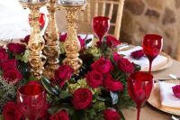 a refined Valentine’s Day wedding centerpiece of greenery, red roses, refined gold candleholders and red glasses to match