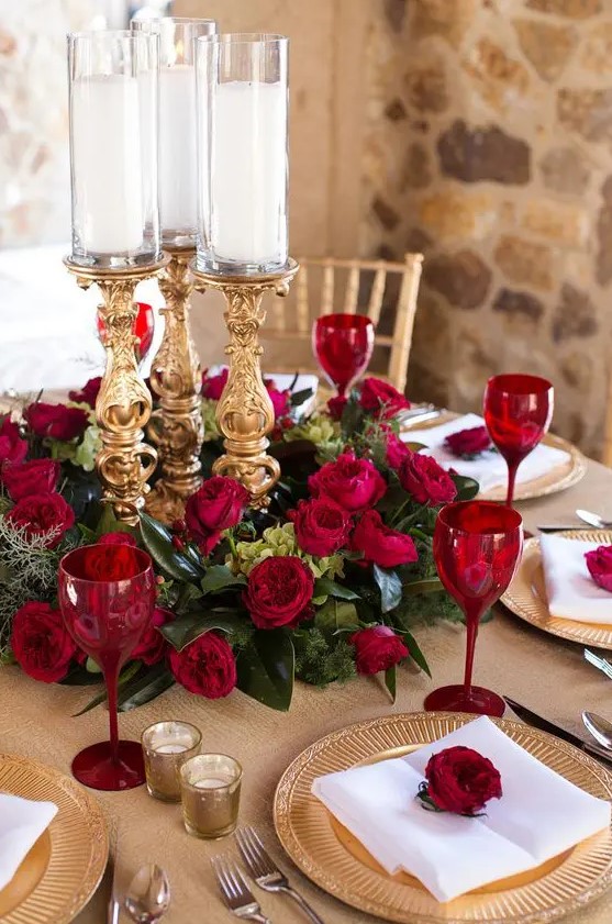 a refined Valentine's Day wedding centerpiece of greenery, red roses, refined gold candleholders and red glasses to match