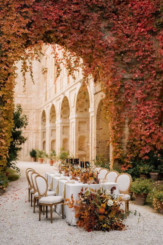 a refined outdoor fall wedding reception space with an arch covered with yellow and red leaves, with matching leaf and bloom arrangements on the table