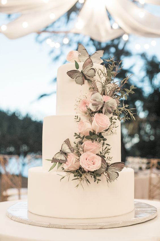 a refined wedding cake in white decorated with greenery, blush roses, grey butterflies is a chic and stylish idea